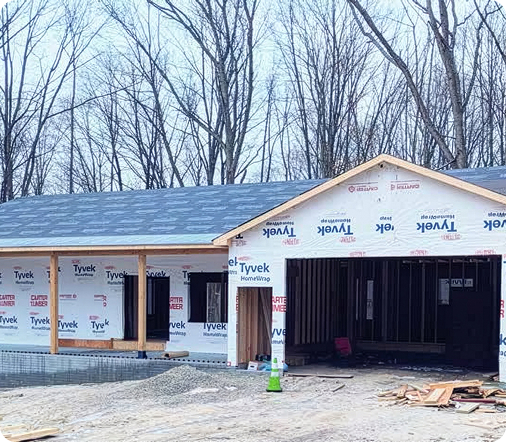 A new house under construction featuring white Tyvek home wrap and a gray shingled roof, showing a housing development.
