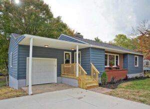 Renovated ranch house with blue siding, a new white garage door, a fresh wooden porch, and professional landscaping.